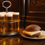 Holztisch in einem Kölner Brauhaus mit mehreren schlanken Kölschgläsern im Kranz, daneben ein Roggenbrötchen mit Käse und Senf, warmes Licht